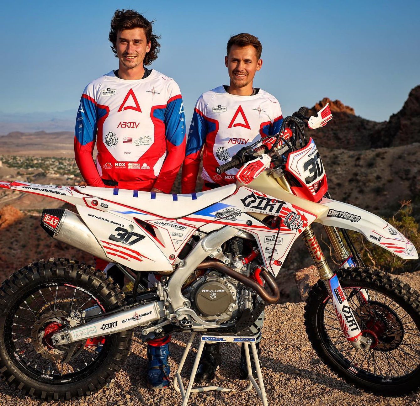 Two team riders behind a race-prepared bike in the desert